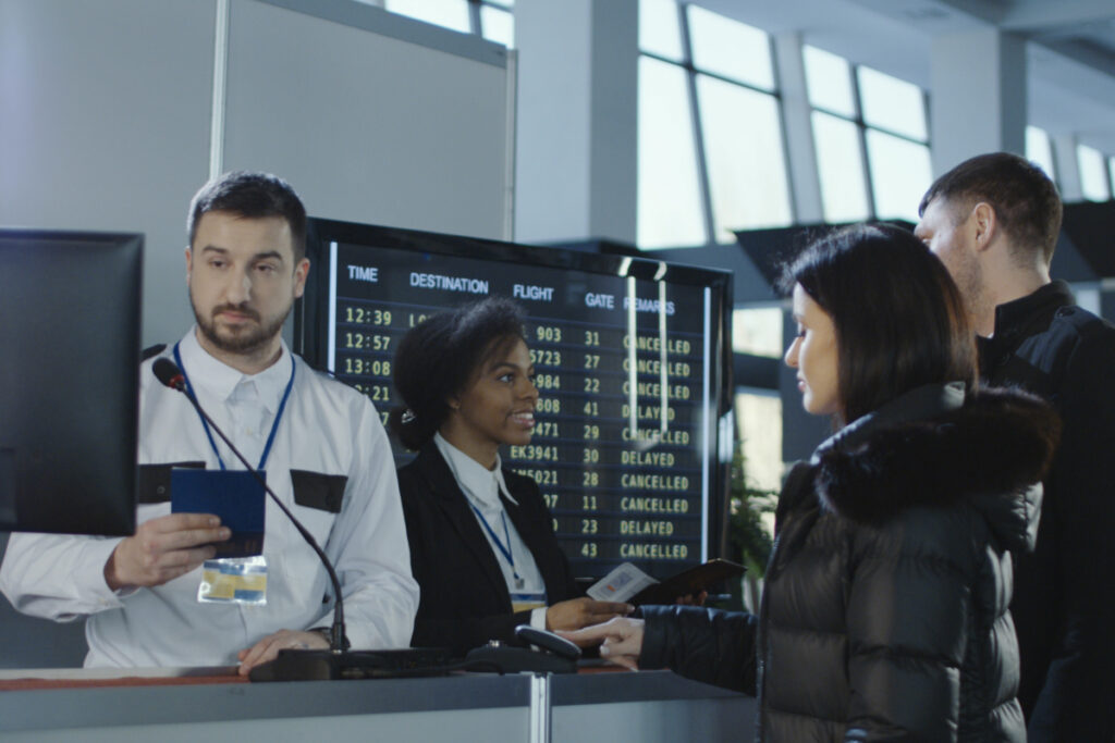 Airport check-in staff assisting passengers at a busy airline desk with departures board showing cancelled and delayed flights