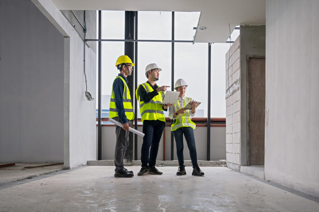 Three construction professionals in hi-vis vests and hard hats reviewing plans on a laptop inside an unfinished building