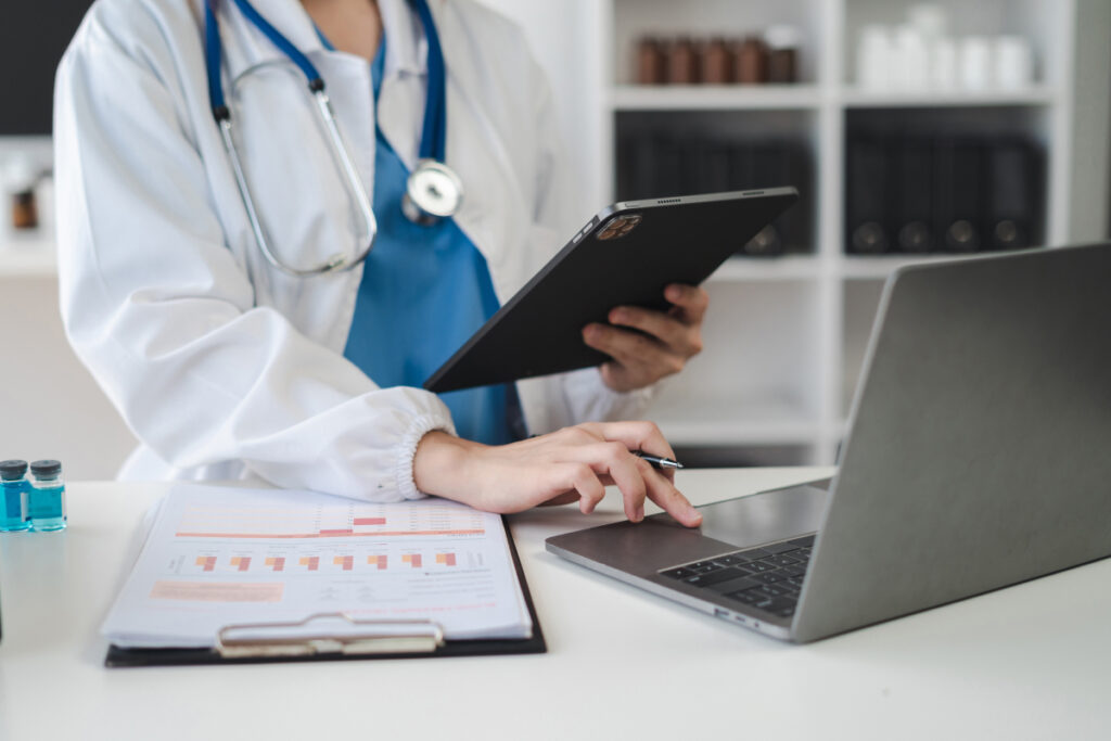 Doctor in white coat and stethoscope reviewing medical charts on a tablet and laptop at a desk