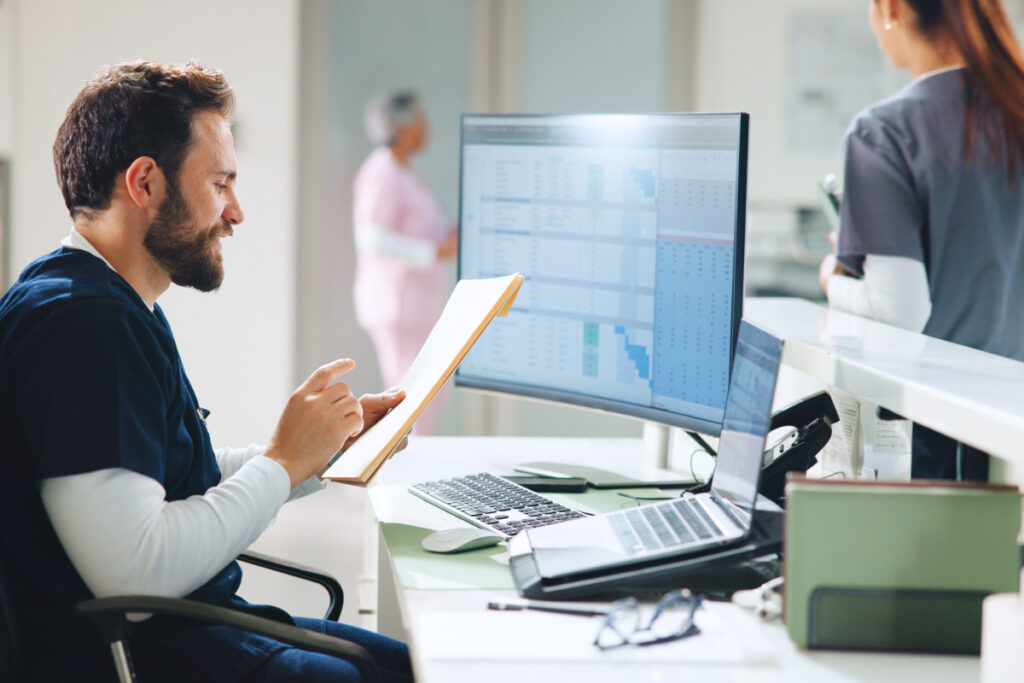 Male nurse or doctor in scrubs reviewing patient records at a hospital reception desk
