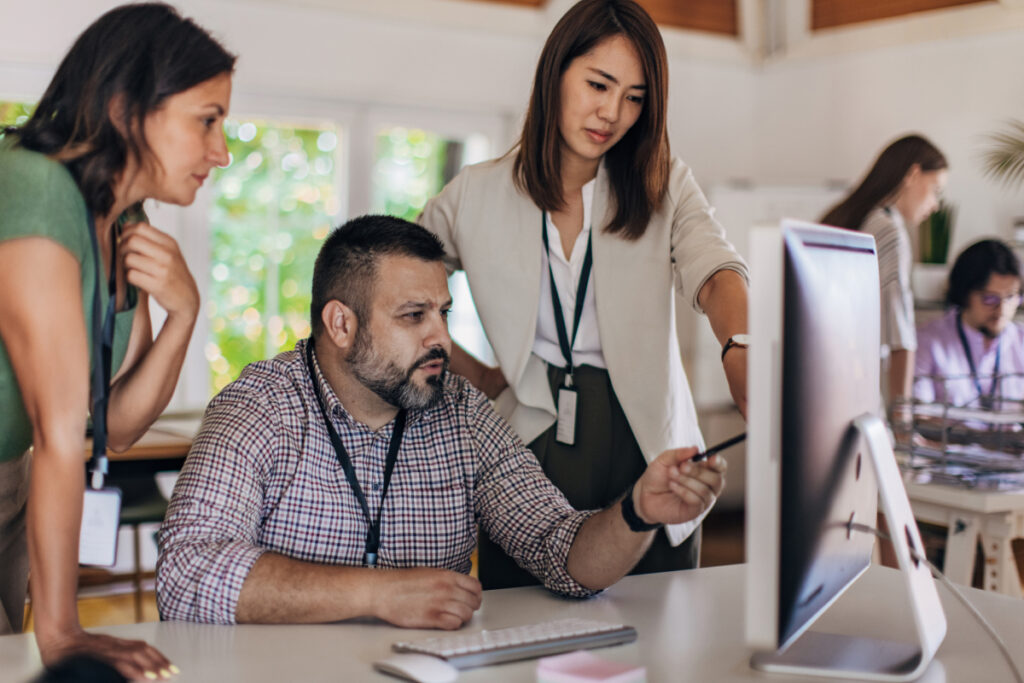 Three office colleagues collaborating around a desktop computer in a bright open-plan workspace