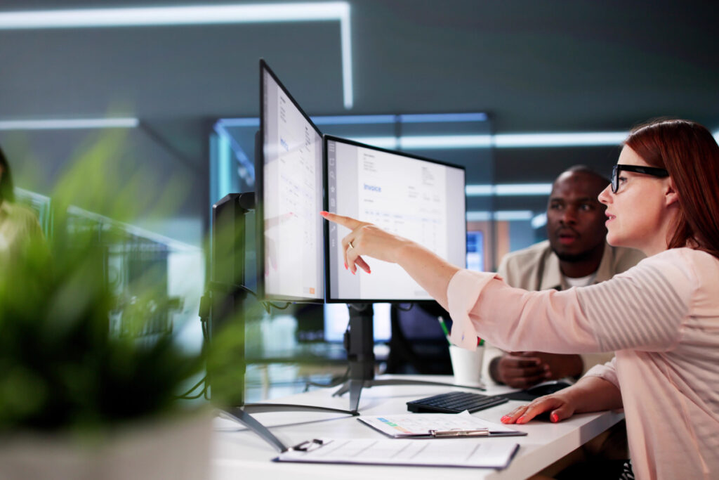 Two office colleagues collaborating at a desk with dual monitors reviewing data and invoices on screen