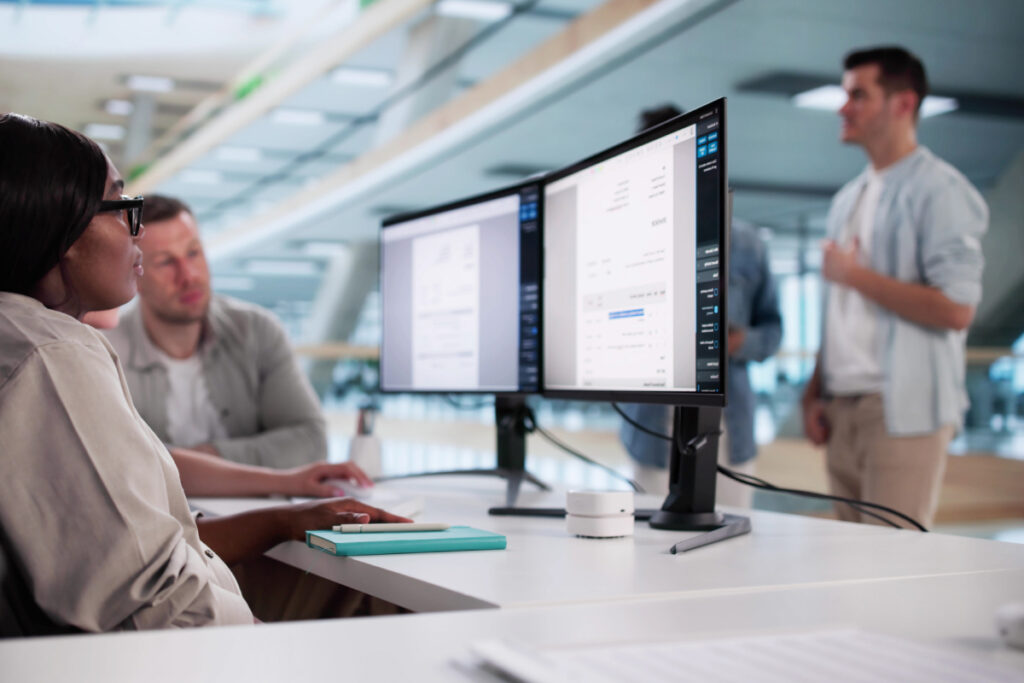 Office team listening to a colleague present while working at a desk with dual monitors displaying software data in a modern open-plan office